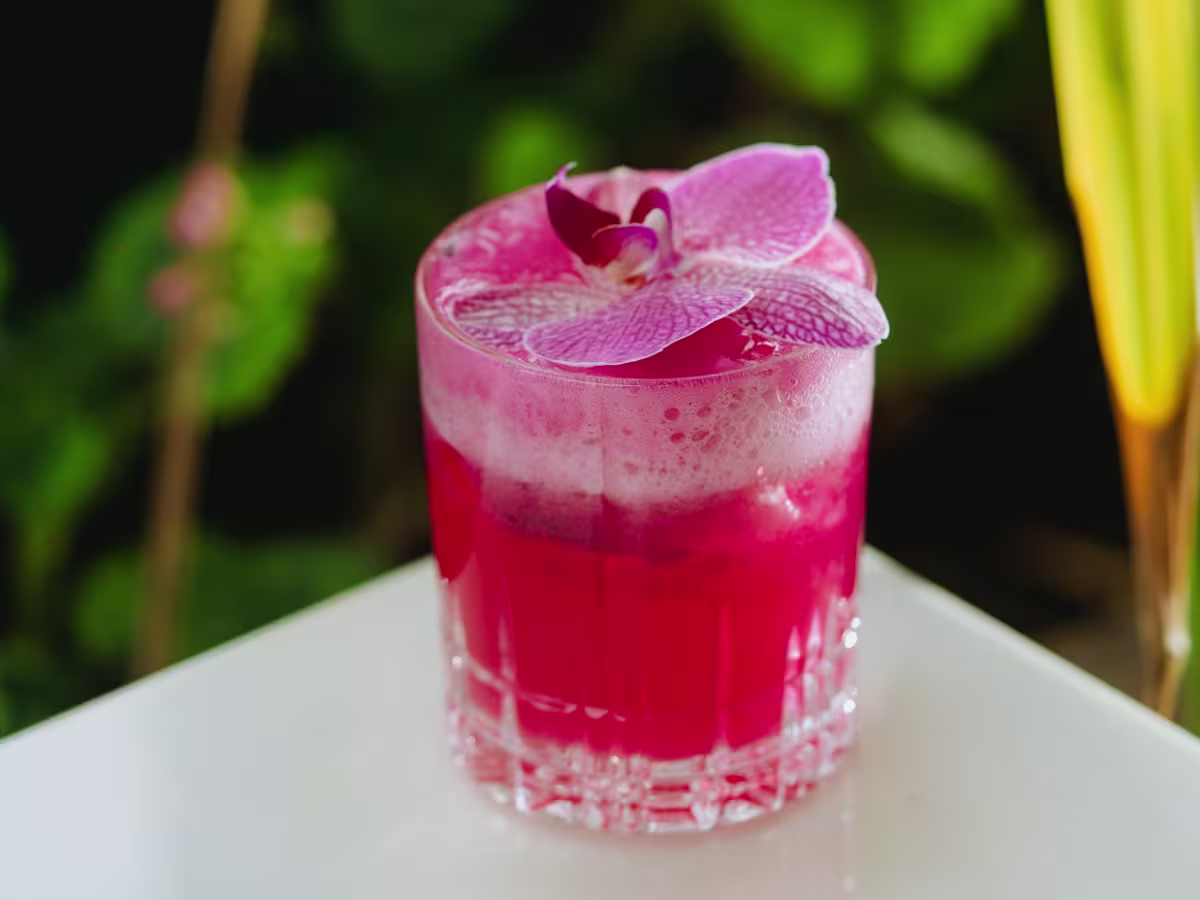 A bright pink cocktail with a frothy top is served in a clear glass and garnished with a purple orchid flower, placed on a white surface with a green background.