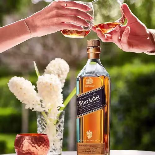Two hands clink whiskey glasses above a bottle of Johnnie Walker Blue Label on a table with white flowers and a copper cup, outdoors.
