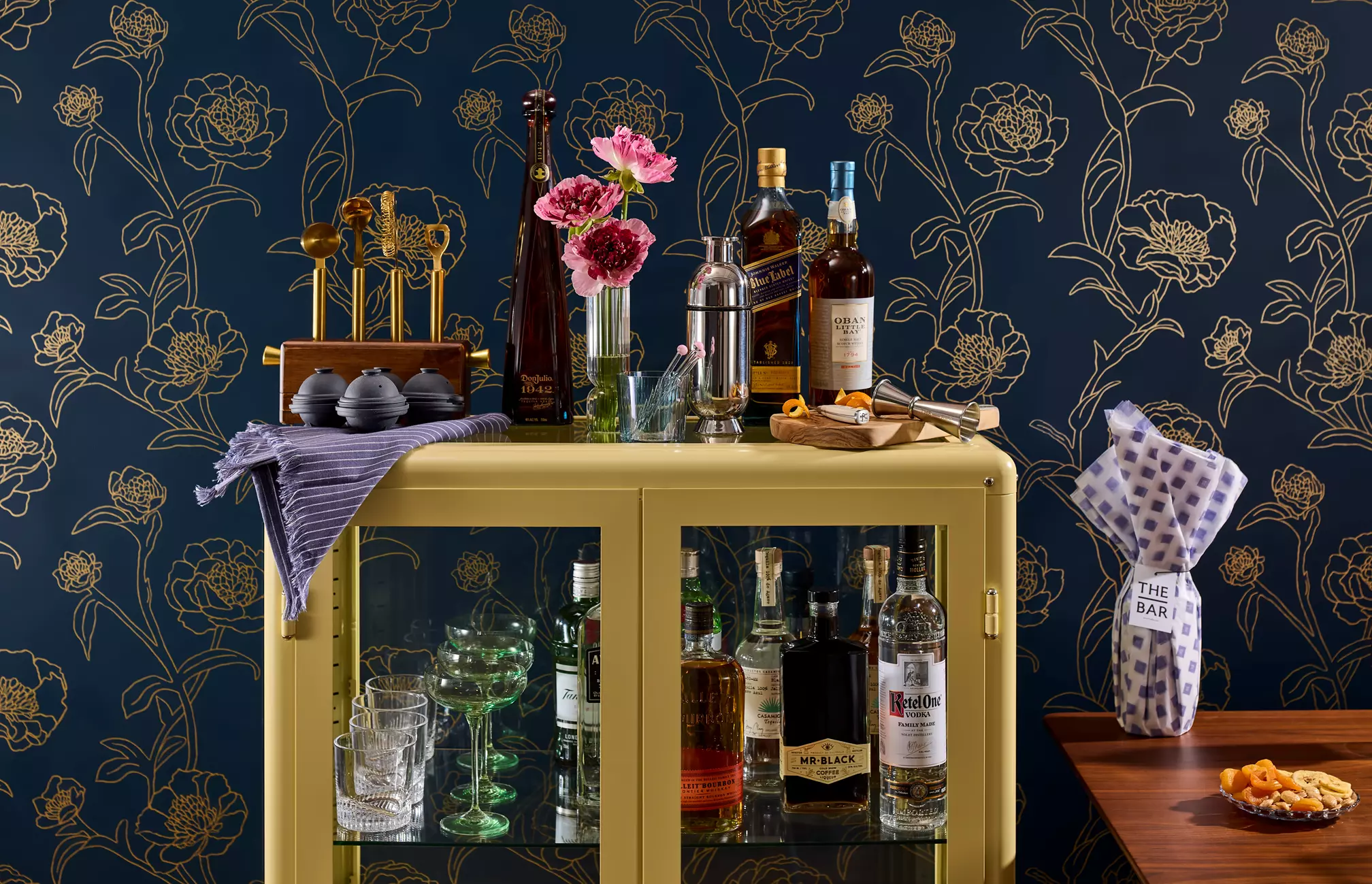 A bar cart with assorted liquor bottles, glassware, bar tools, and pink flowers stands against a dark blue floral wallpaper. A wrapped wine bottle and snack plate sit on a nearby table.