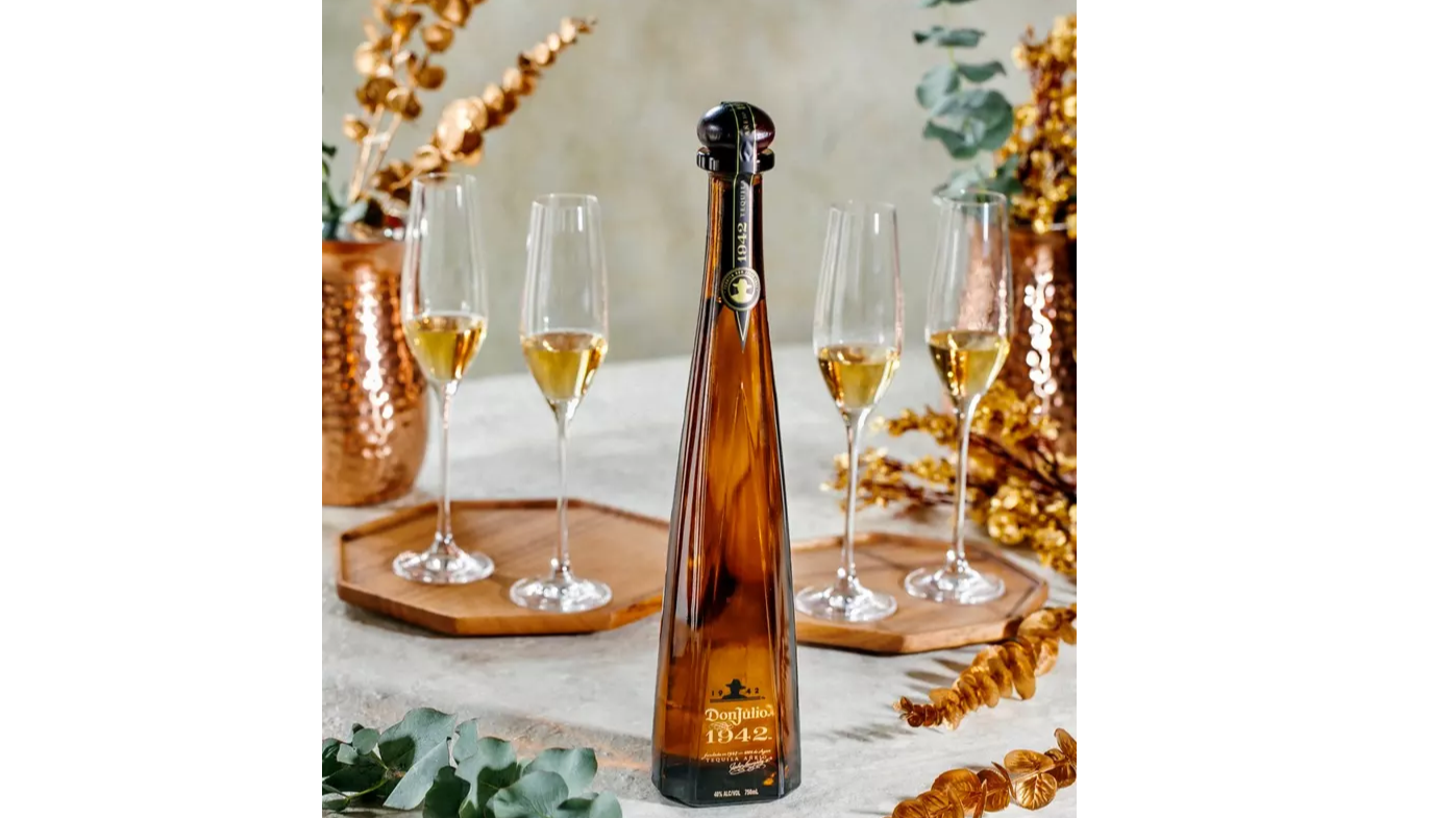 A bottle of Don Julio 1942 tequila is displayed on a table with three filled champagne flutes, copper vases, and decorative greenery and golden branches.