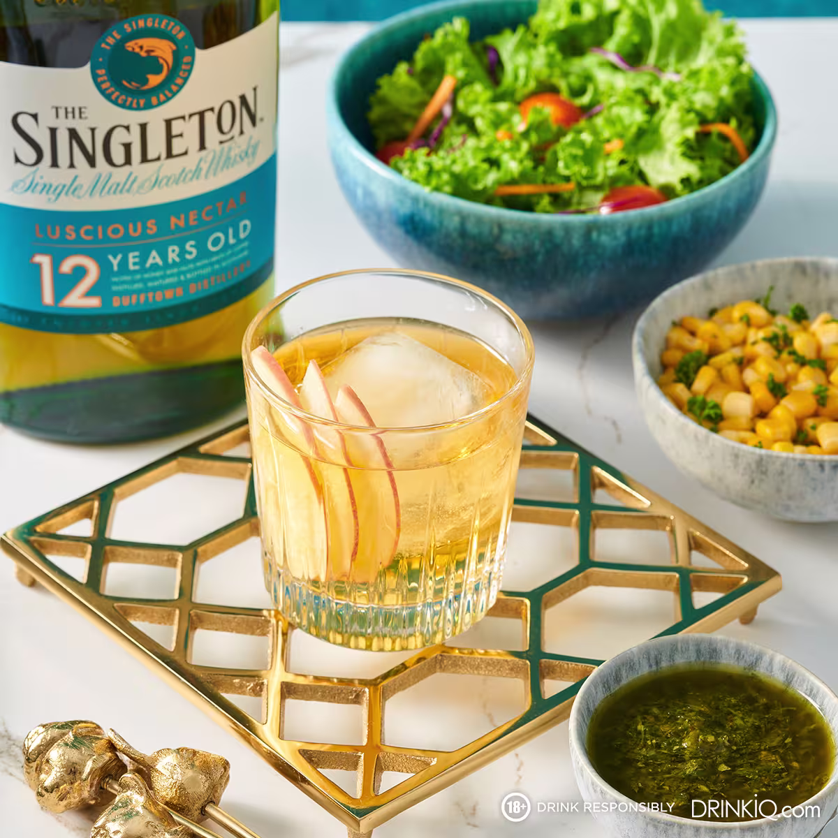 The Singleton whisky cocktail in a coupe glass garnished with fresh herbs, styled on a gold geometric tray alongside a salad, shot on a mint green background