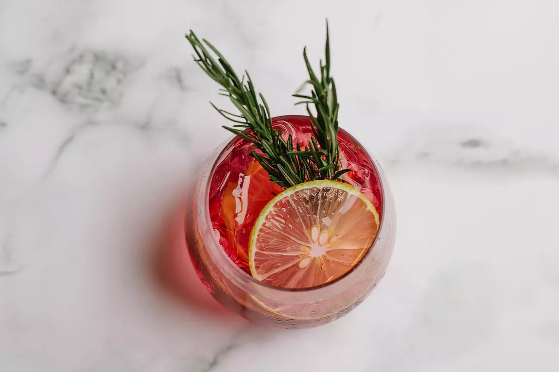 A glass of red cocktail with ice, garnished with a slice of lemon and a sprig of rosemary, on a white marble surface.