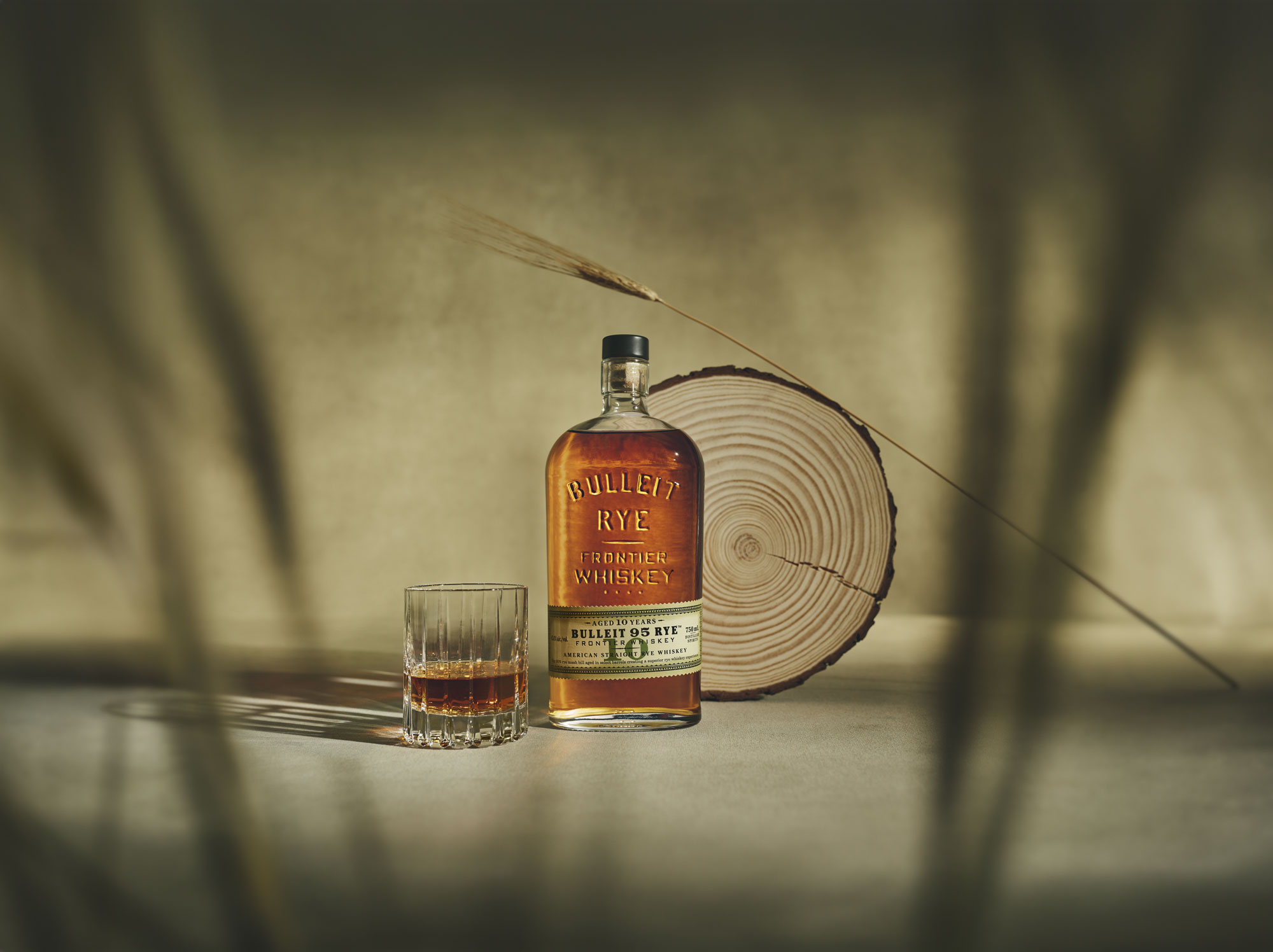A bottle of Bulleit Rye Frontier Whiskey and a glass with whiskey sit on a surface, beside a wood slice and a dried wheat stalk, with blurred foreground elements.