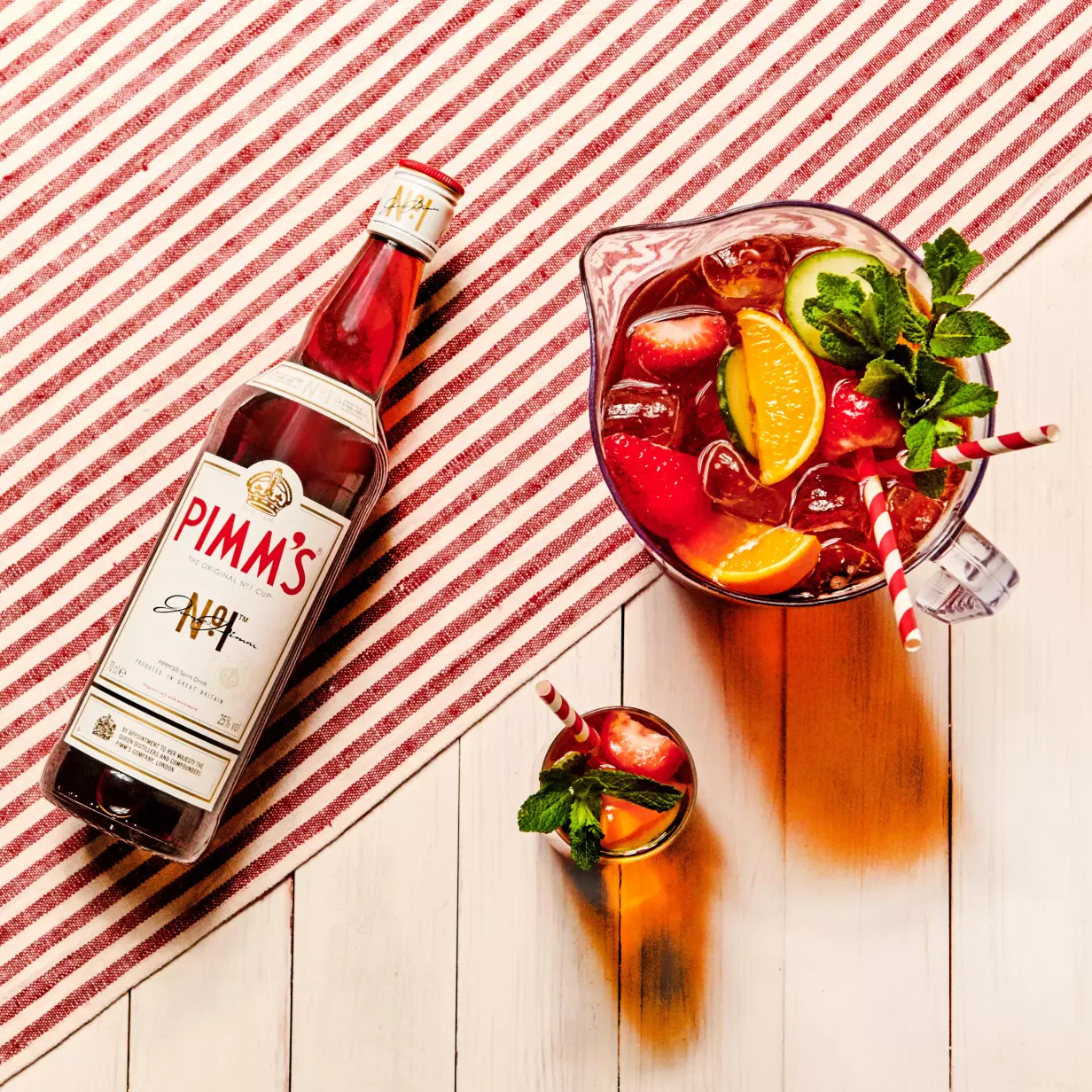A bottle of Pimms, a glass, and a pitcher filled with iced Pimms cocktail, citrus slices, strawberries, and mint sit on a white wooden table with a red-striped cloth.