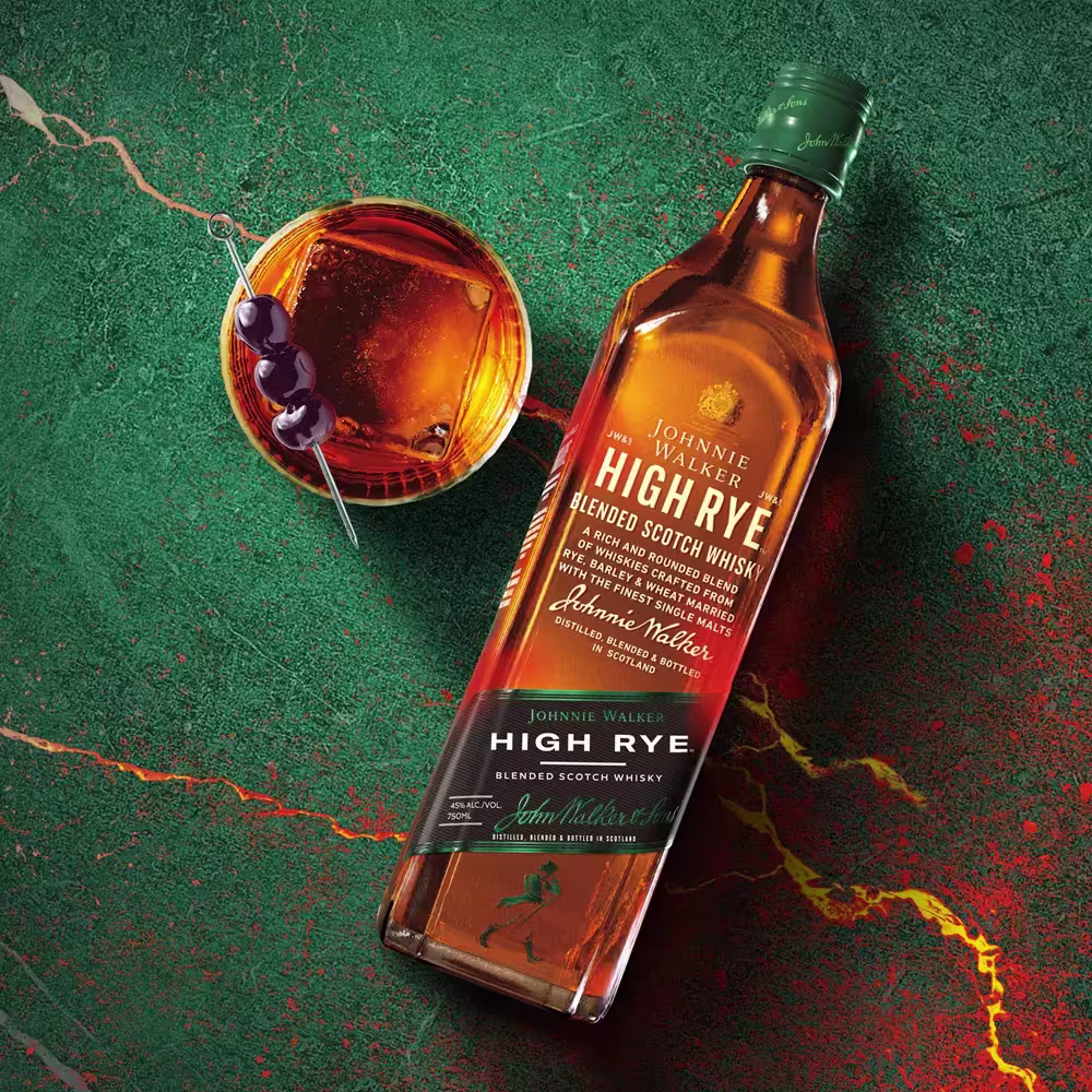 A bottle of Johnnie Walker High Rye Blended Scotch Whisky lies next to a cocktail garnished with three cherries on a green and red marble surface.