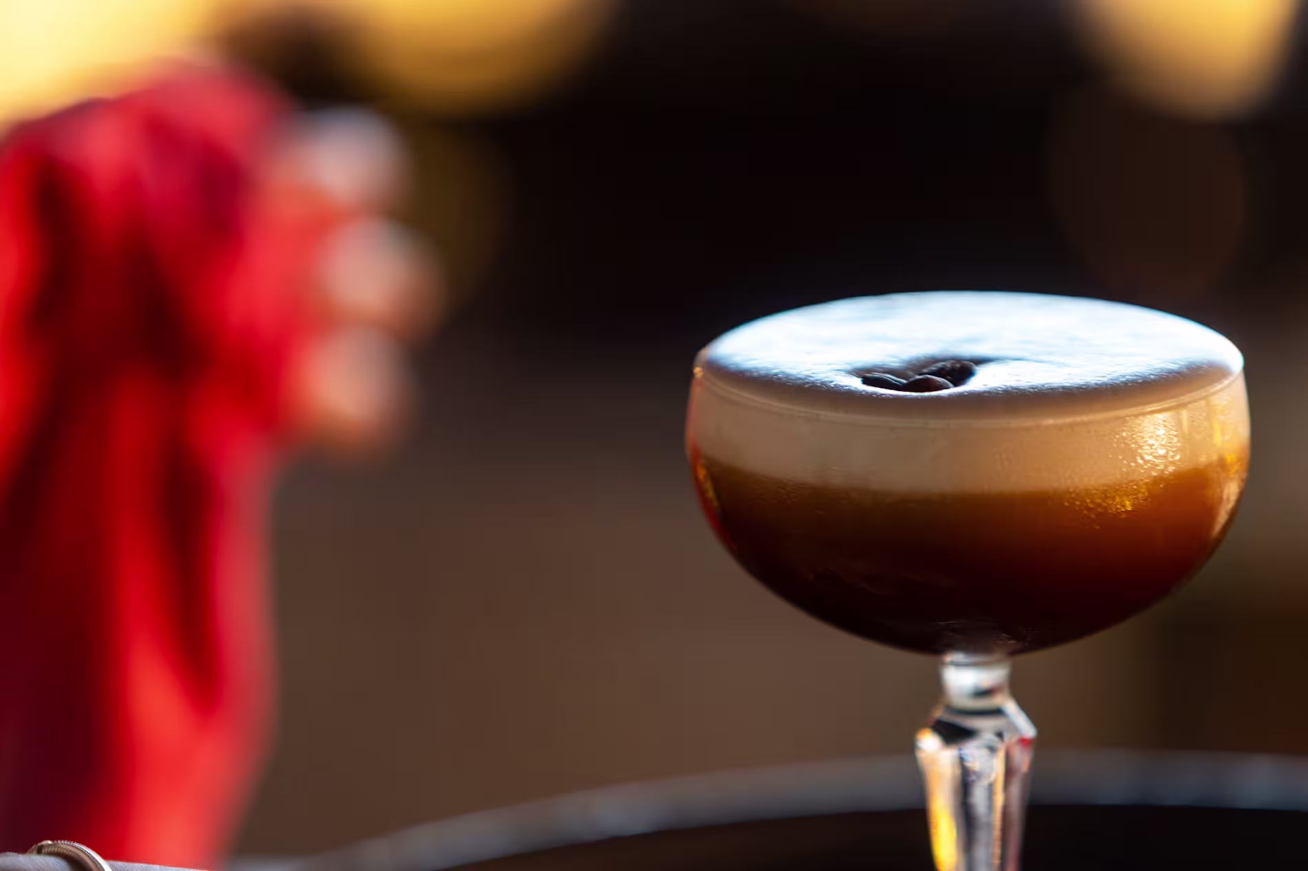 Cóctel de café en copa elegante con espuma cremosa y un grano de café decorativo, iluminado con luz cálida y fondo desenfocado.