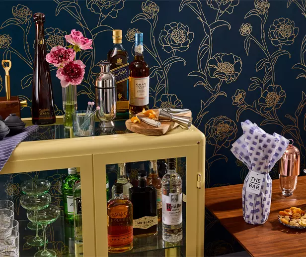 Home bar setup with various liquor bottles, glassware, a cocktail shaker, snacks, and a gift-wrapped bottle, set against a dark floral wallpaper background.