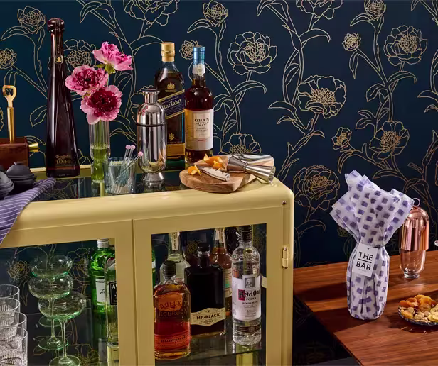 Home bar setup with various liquor bottles, glassware, a cocktail shaker, snacks, and a gift-wrapped bottle, set against a dark floral wallpaper background.
