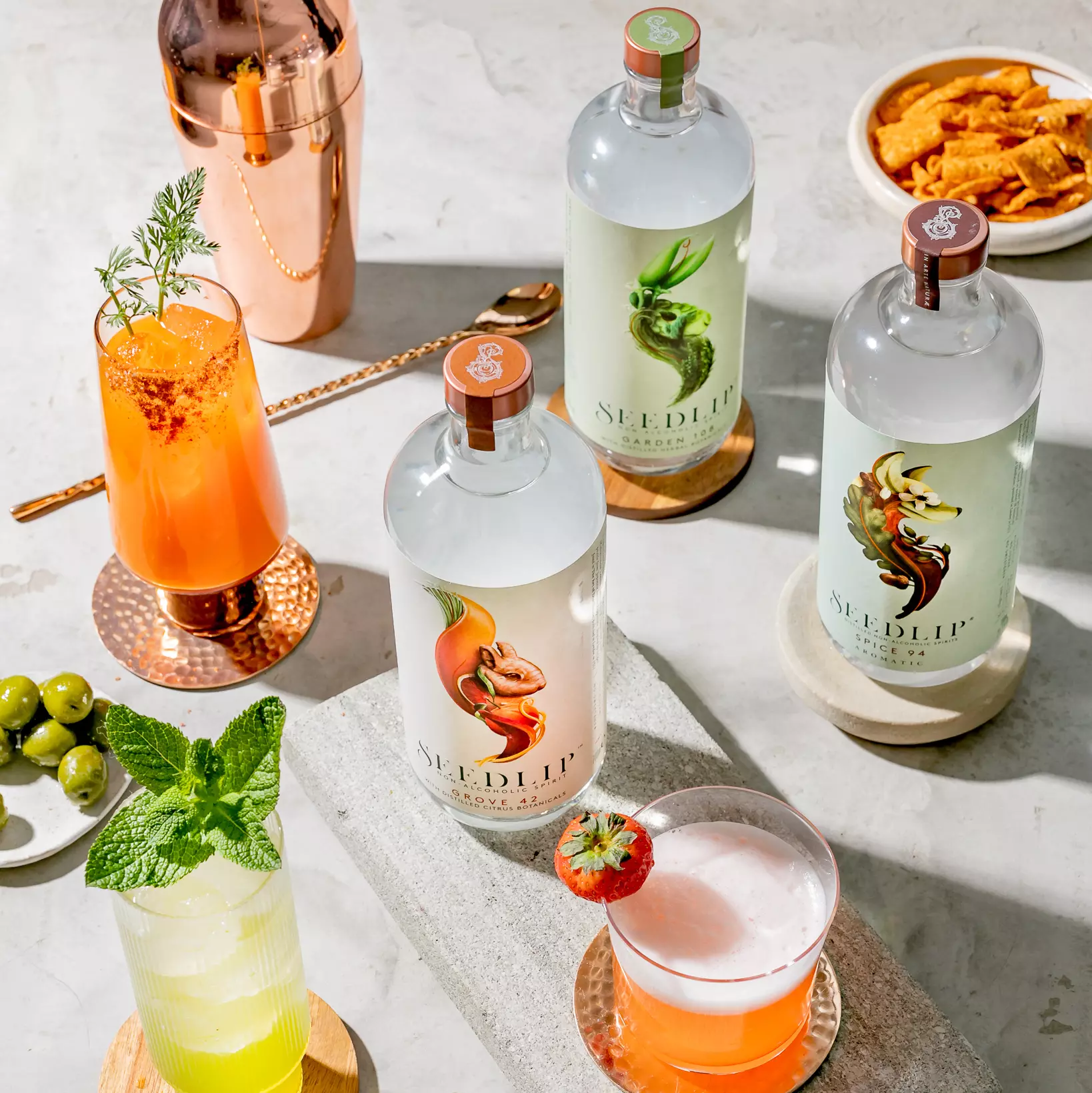 Three bottles of Seedlip non-alcoholic spirits are arranged on a table with cocktails, garnishes, green olives, snacks, and a copper cocktail shaker.