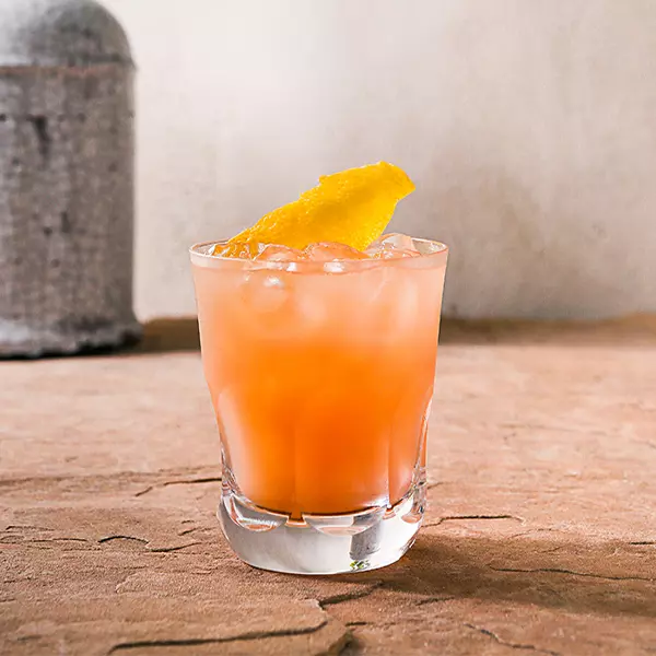 A short glass filled with an orange-colored iced drink, garnished with a lemon peel, sits on a textured brown surface against a plain background.