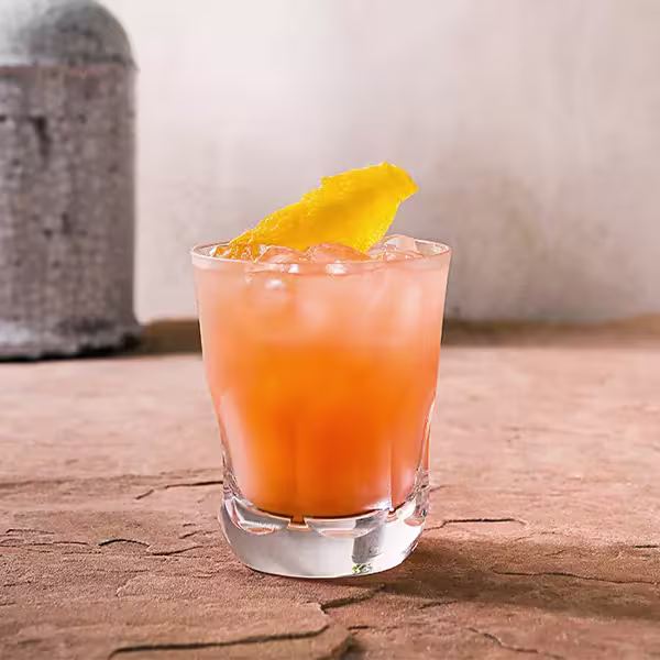 A short glass filled with an orange-colored iced drink, garnished with a lemon peel, sits on a textured brown surface against a plain background.