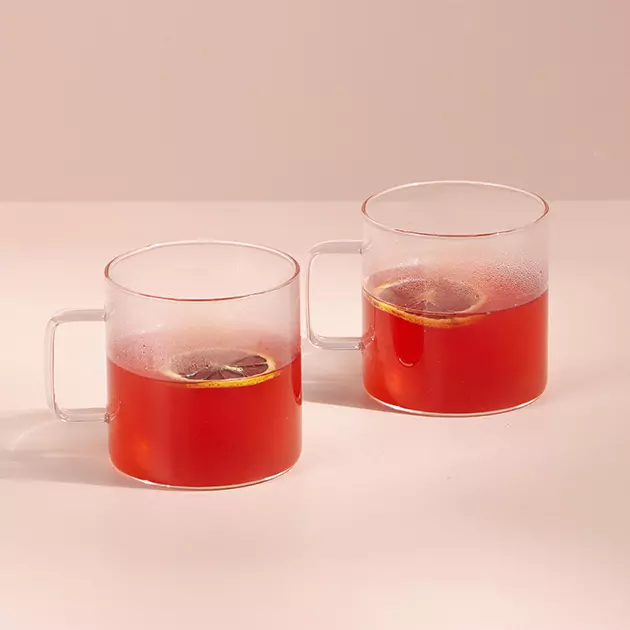 Two clear glass mugs filled with red tea, each with a lemon slice, placed on a light surface against a plain background.