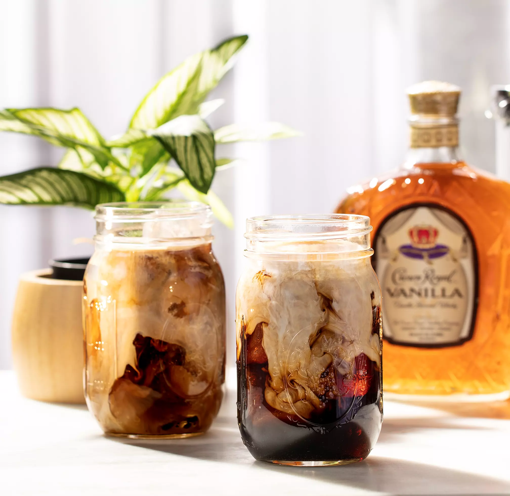 Two mason jars filled with iced coffee and cream sit on a table beside a bottle of vanilla whiskey and a potted plant.
