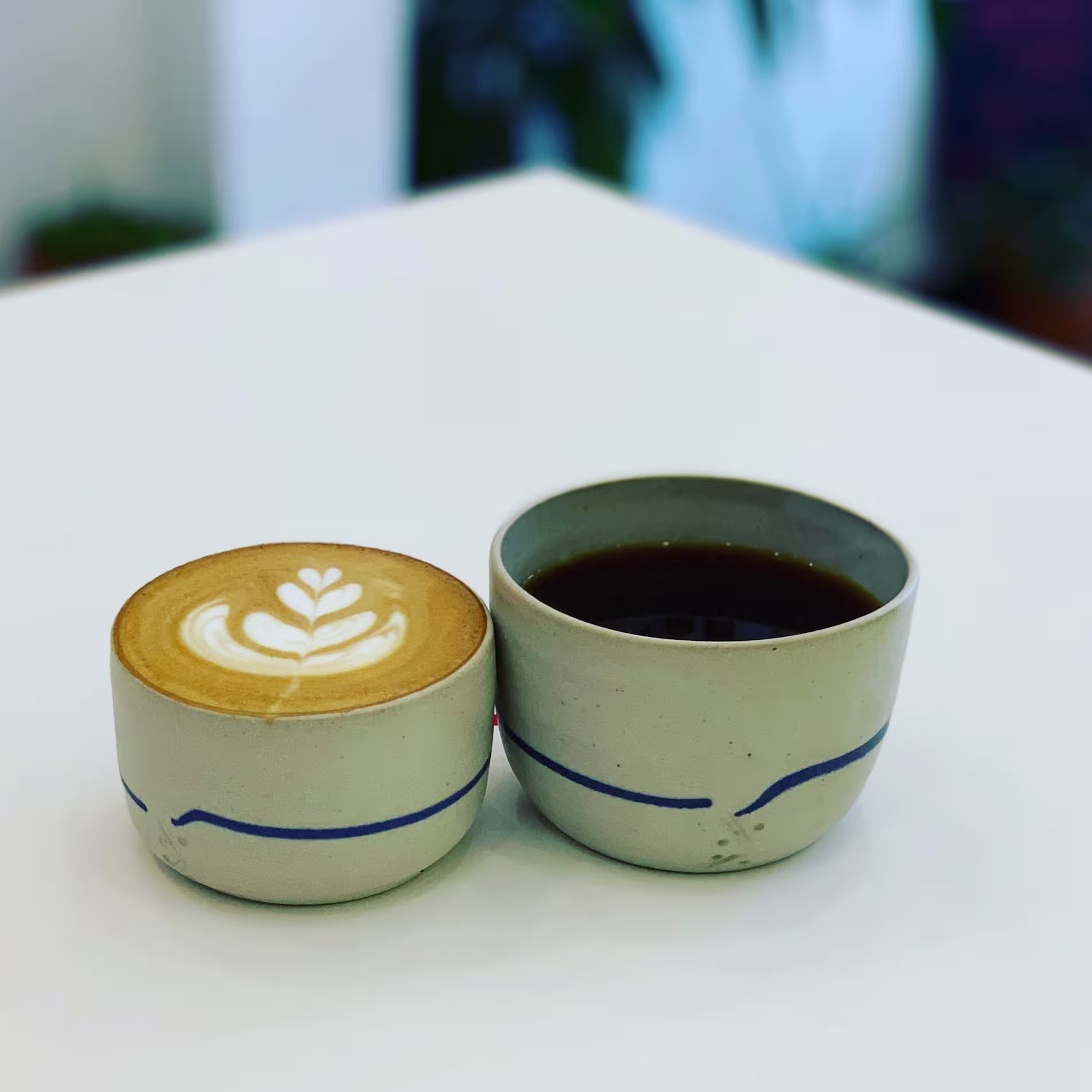 Dos tazas de cerámica en Parcería, una con latte art en forma de hoja y otra con café solo, sobre una mesa blanca minimalista.