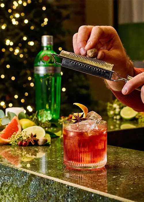A person grates nutmeg over a red cocktail with ice and a citrus garnish on a bar, with a green bottle and assorted fruits in the background.