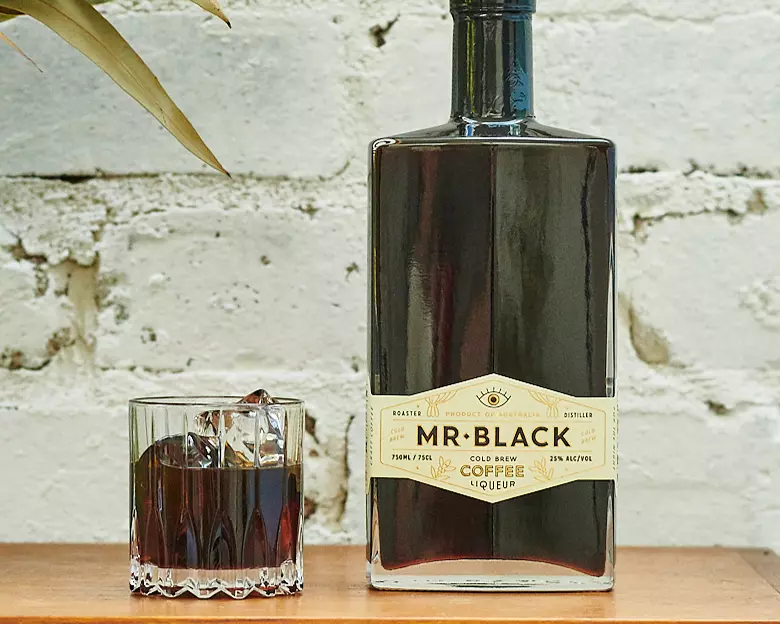 A bottle of Mr. Black Cold Brew Coffee Liqueur stands next to a glass of iced coffee liqueur on a wooden surface against a white brick wall.