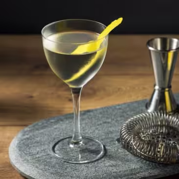 A martini in a stemmed glass garnished with a lemon twist, sitting on a bar surface next to a jigger, a strainer, and a whole lemon.