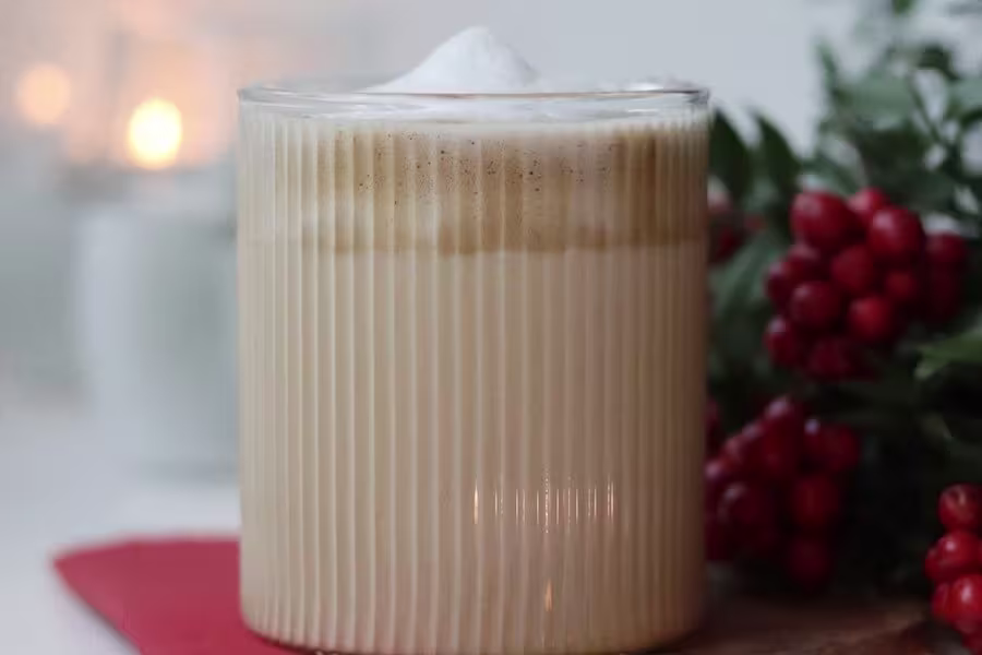 A glass of layered latte with foam on top sits on a wooden surface, with red berries and greenery in the background.