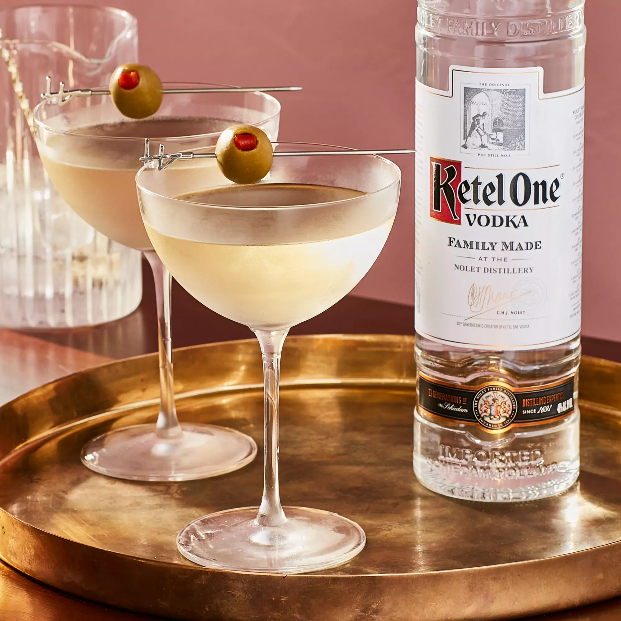 Two martini glasses garnished with olives on a gold tray next to a tall bottle of Ketel One vodka against a pink background.