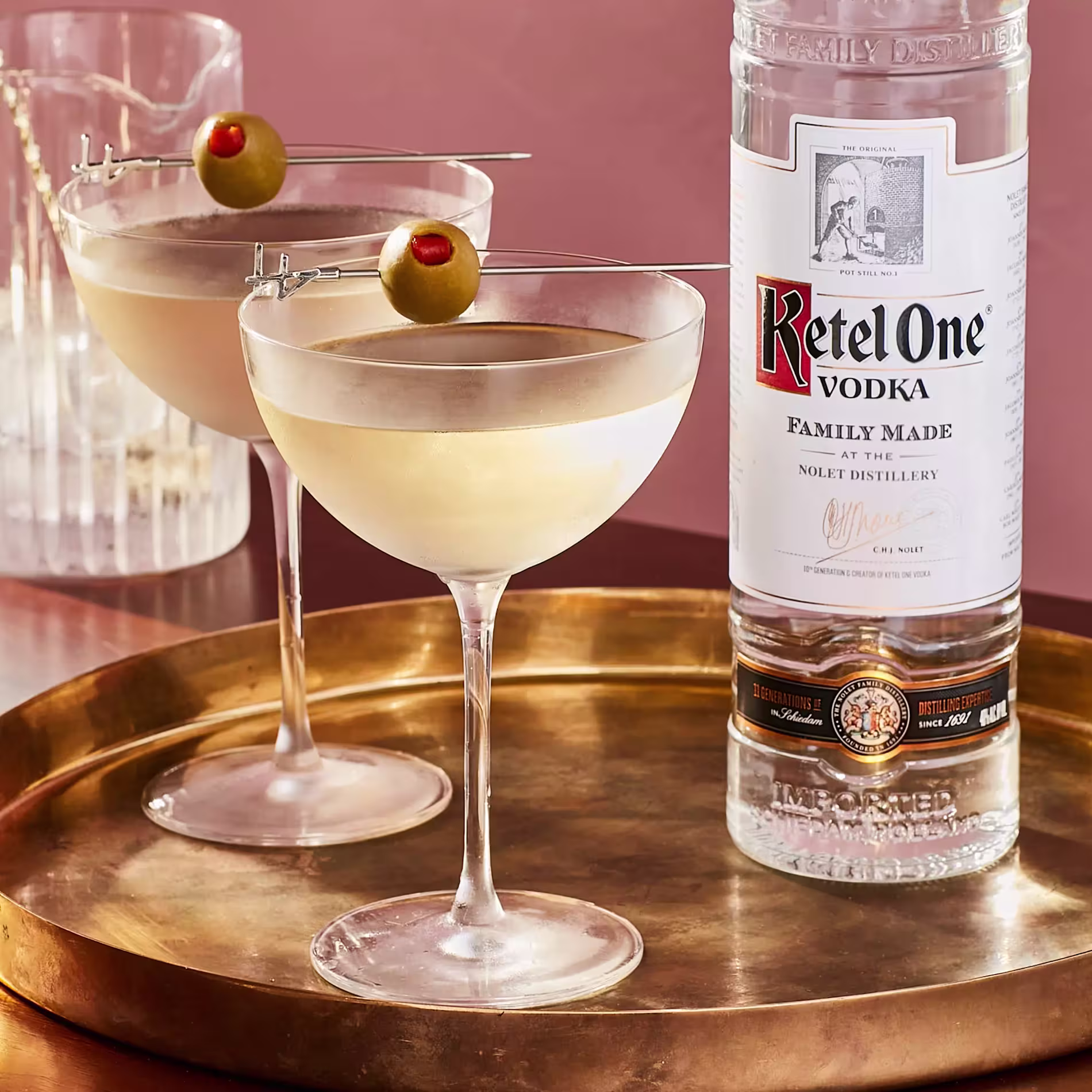 Two martini glasses garnished with olives on a gold tray next to a tall bottle of Ketel One vodka against a pink background.