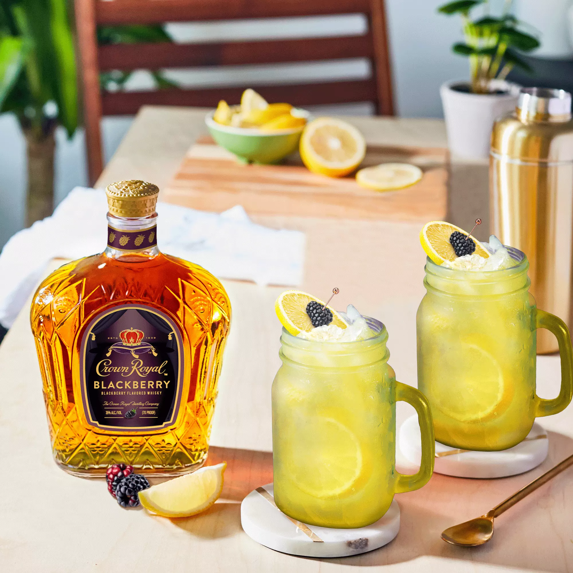 A bottle of Crown Royal Blackberry whiskey sits next to two mason jar cocktails garnished with lemon, whipped cream, and a blackberry on a wooden table with fresh fruit.