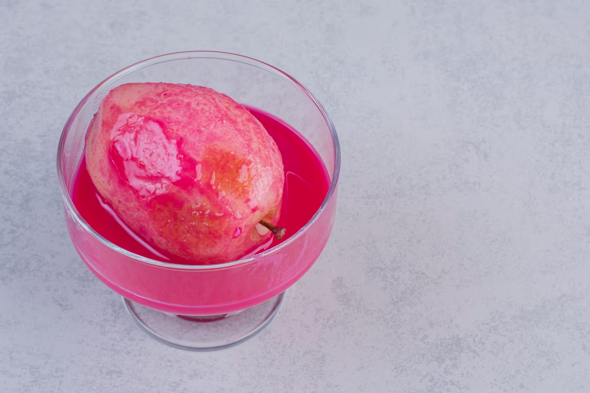A whole guava fruit is placed in a glass filled with bright pink liquid, set on a light gray textured surface.