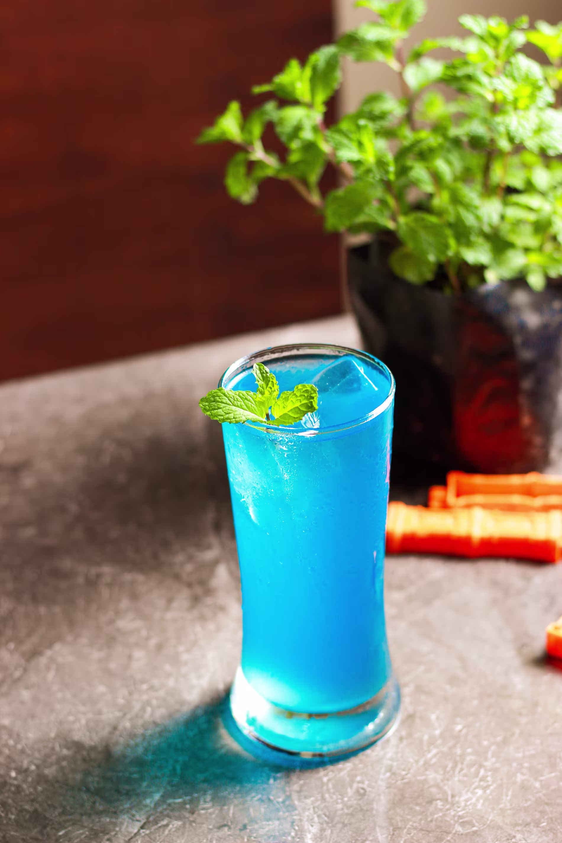 A tall glass of blue beverage with ice cubes and a mint garnish sits on a gray surface, with a potted plant and orange straws in the background.