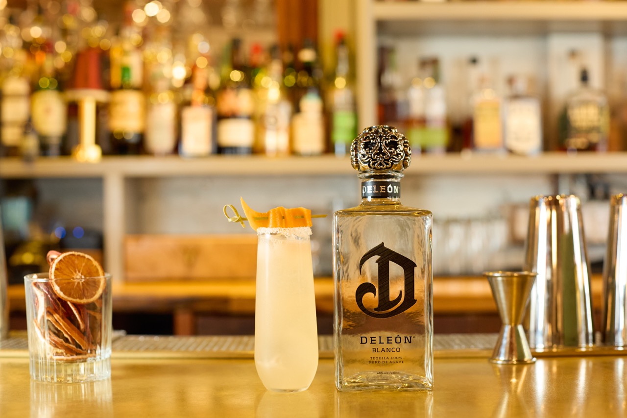 A bottle of DeLeón Blanco tequila, a cocktail garnished with orange peel, a glass with dried citrus, and bar tools on a bar counter with liquor bottles in the background.