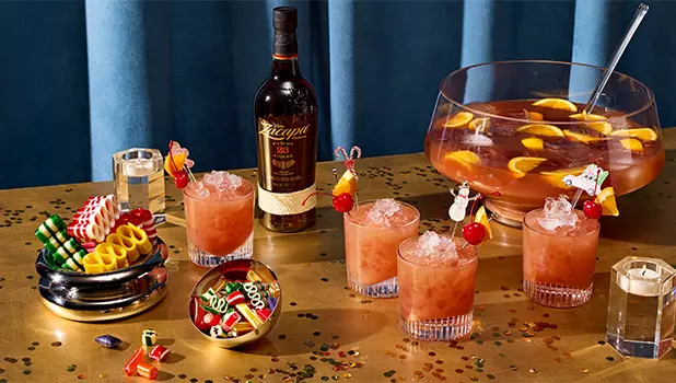 A bottle of rum, four cocktails with fruit garnishes, a punch bowl with orange slices, snacks, dice, and confetti on a gold table with blue background.