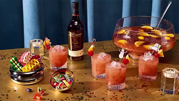 A bottle of rum, four cocktails with fruit garnishes, a punch bowl with orange slices, snacks, dice, and confetti on a gold table with blue background.