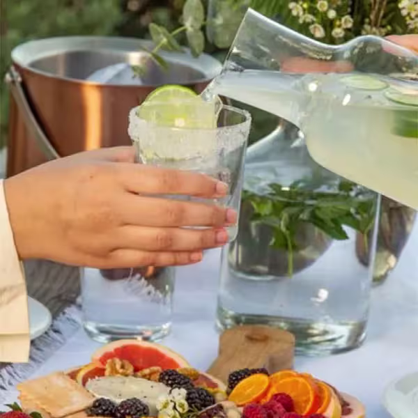A person pours a clear beverage with lime from a glass pitcher into a glass with a salted rim, next to a charcuterie board with fruit.