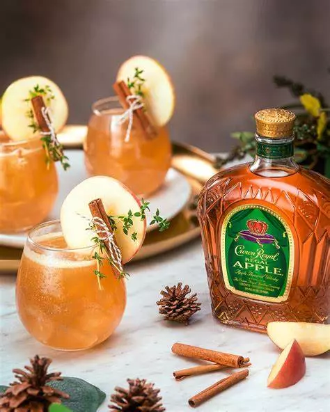 Three glasses of apple cocktails garnished with apple slices, cinnamon sticks, and herbs sit beside a bottle of Crown Royal Regal Apple whisky, with pinecones and cinnamon sticks on the table.
