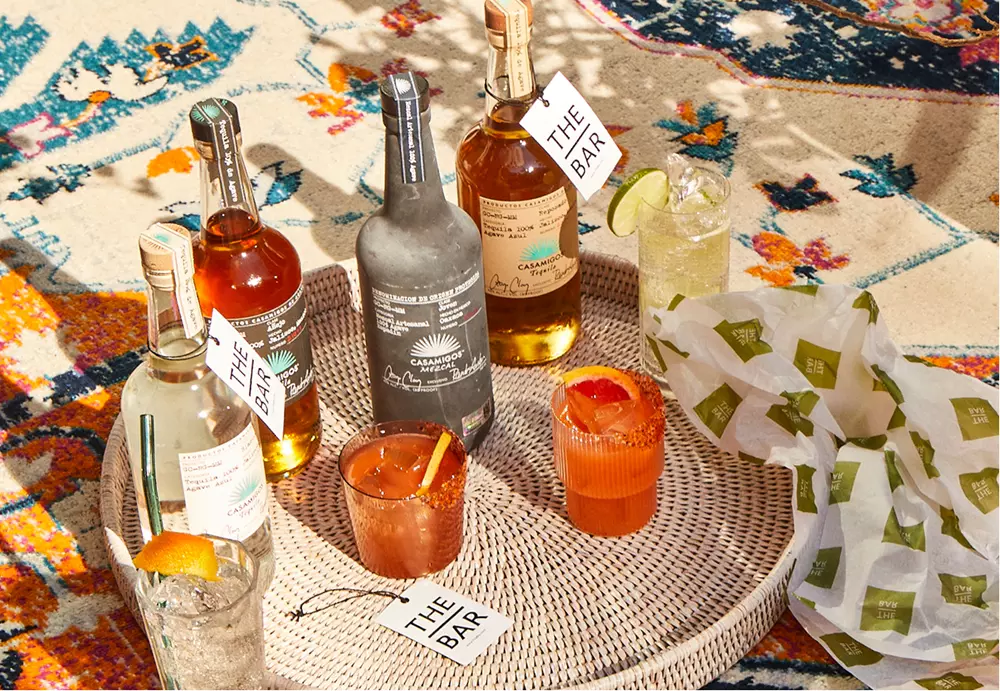 A woven tray on a patterned rug holds five liquor bottles, three cocktails with garnishes, drink tags labeled THE BAR, and a wrapped food item.