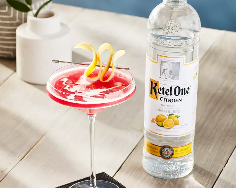 A pink cocktail with a lemon twist garnish in a coupe glass sits next to a bottle of Ketel One Citroen vodka on a wooden table.