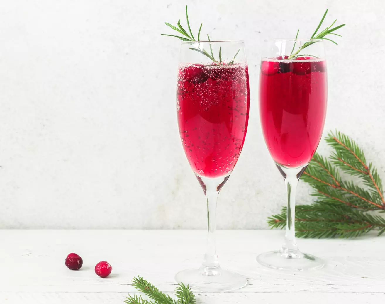 Two champagne flutes filled with red sparkling drink, garnished with cranberries and rosemary sprigs, on a white surface with evergreen branches.