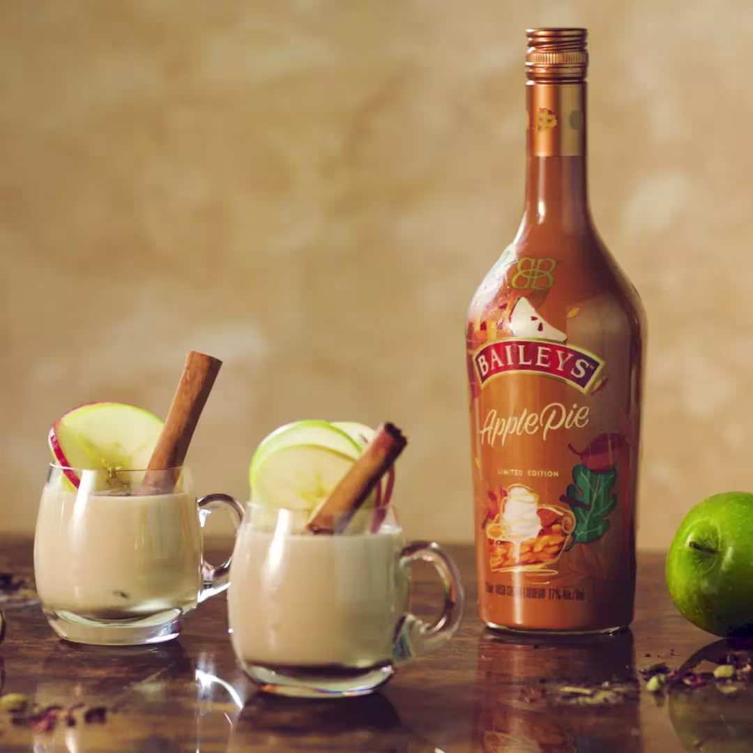 A bottle of Baileys Apple Pie liqueur stands beside two glass mugs filled with a creamy drink, garnished with apple slices and cinnamon sticks; a green apple lies nearby.