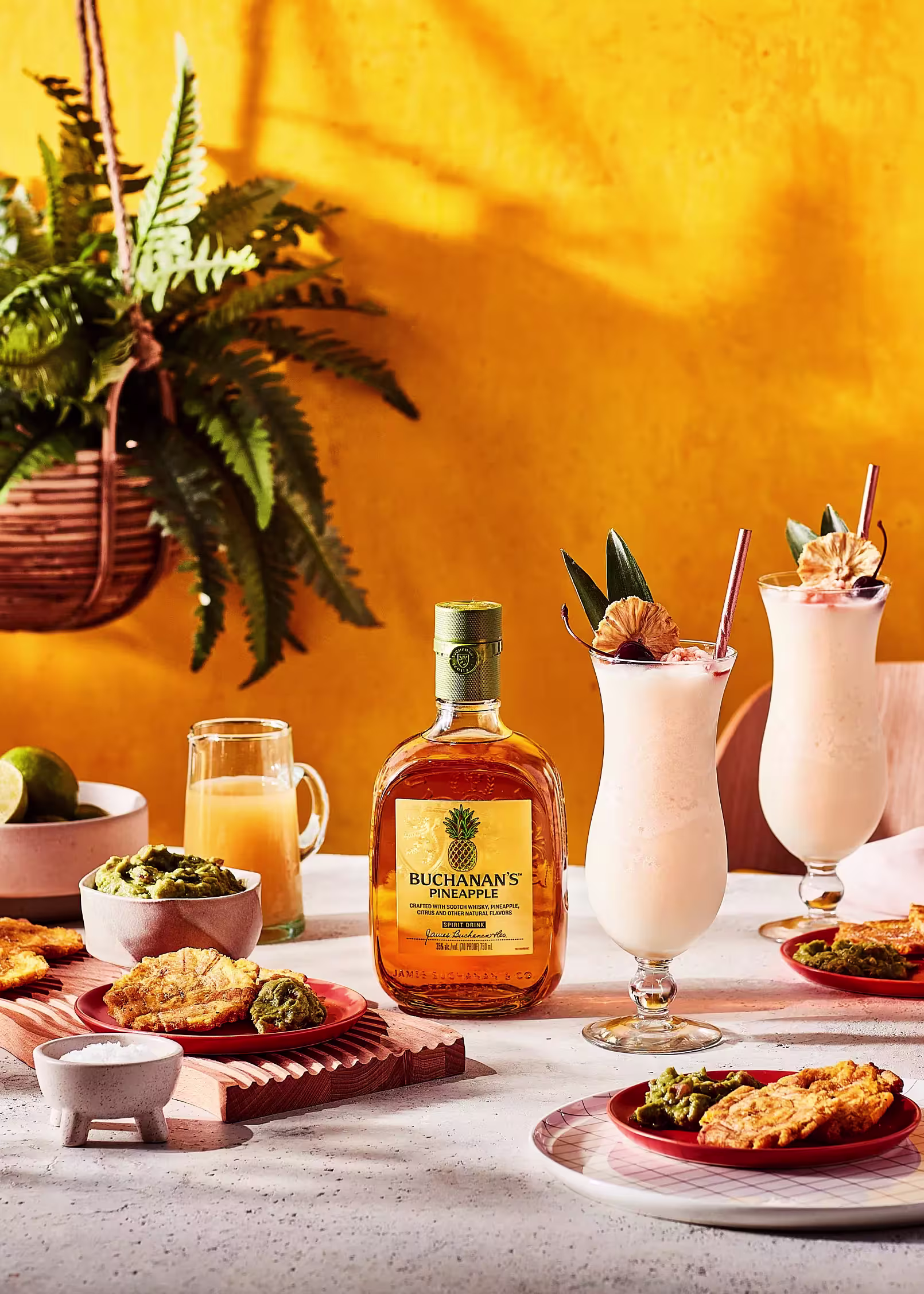A bottle of Buchanan’s Pineapple sits on a table with tropical cocktails, fried snacks with guacamole, and a plant in the background against a bright yellow wall.