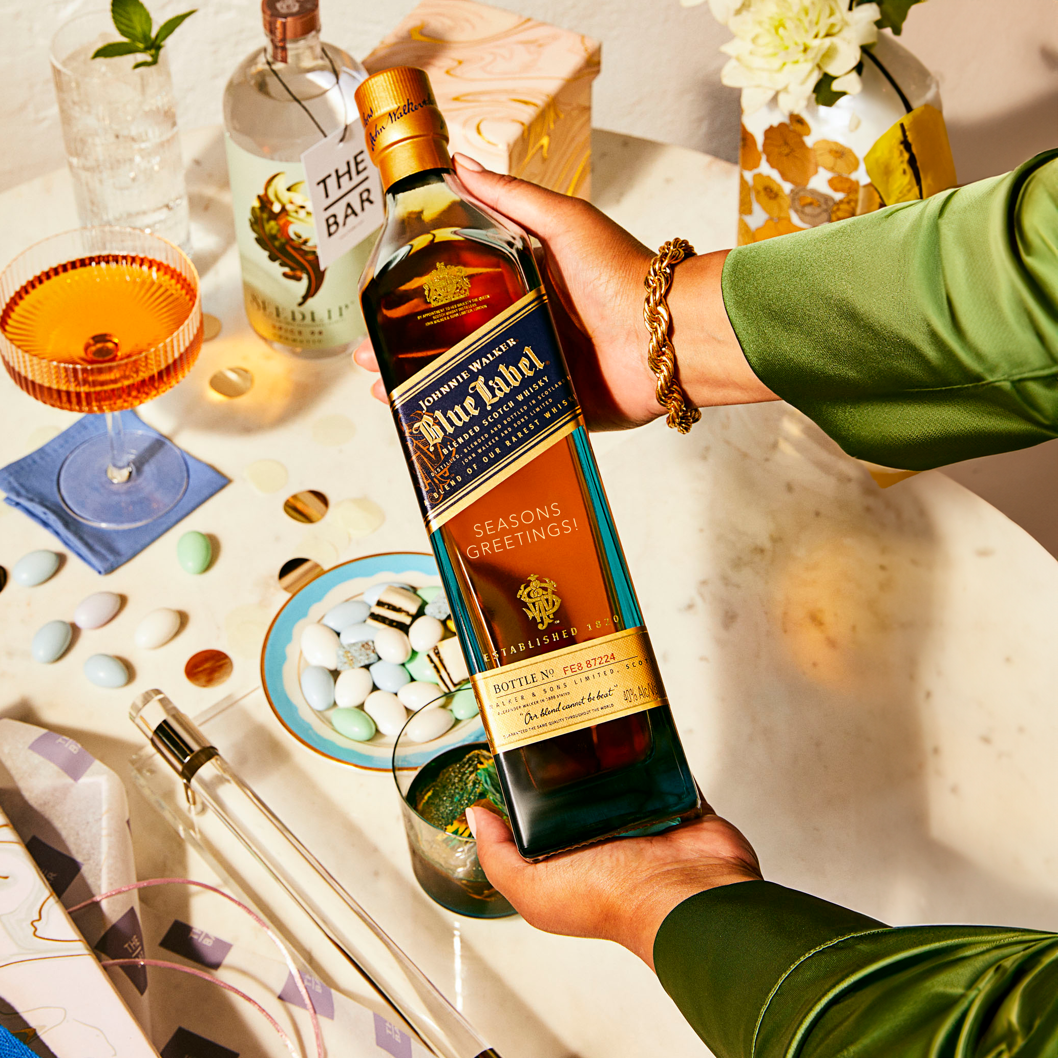 A person holding a bottle of Johnnie Walker Blue Label Seasons Greetings whisky over a table set with drinks, flowers, snacks, and glassware.