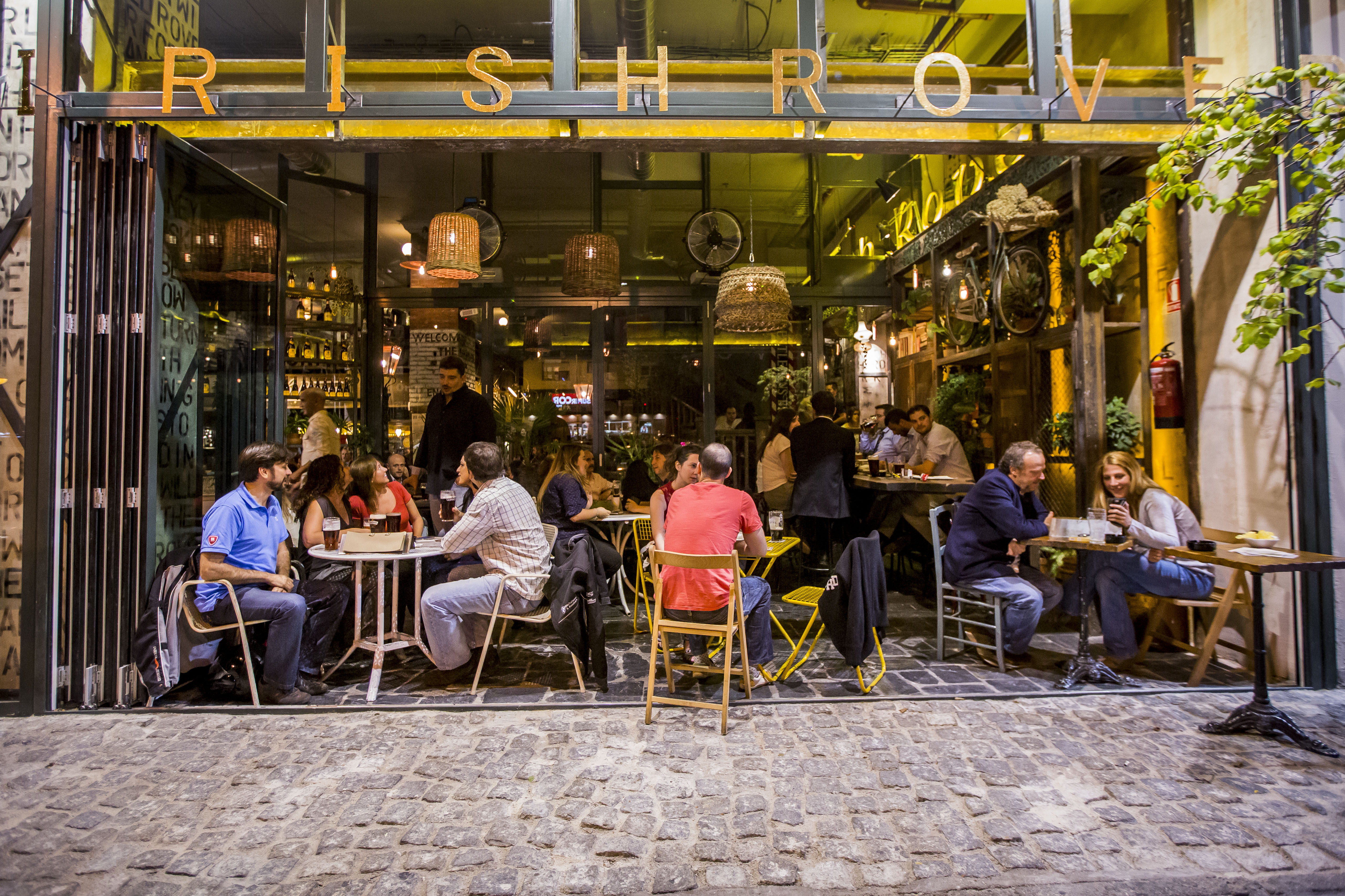 La imagen muestra una zona de asientos al aire libre de el Irish Rover con clientes sentados en varias mesas. Hay luces colgantes y plantas, creando un ambiente acogedor. El letrero sobre la entrada dice “IRISH ROVER”