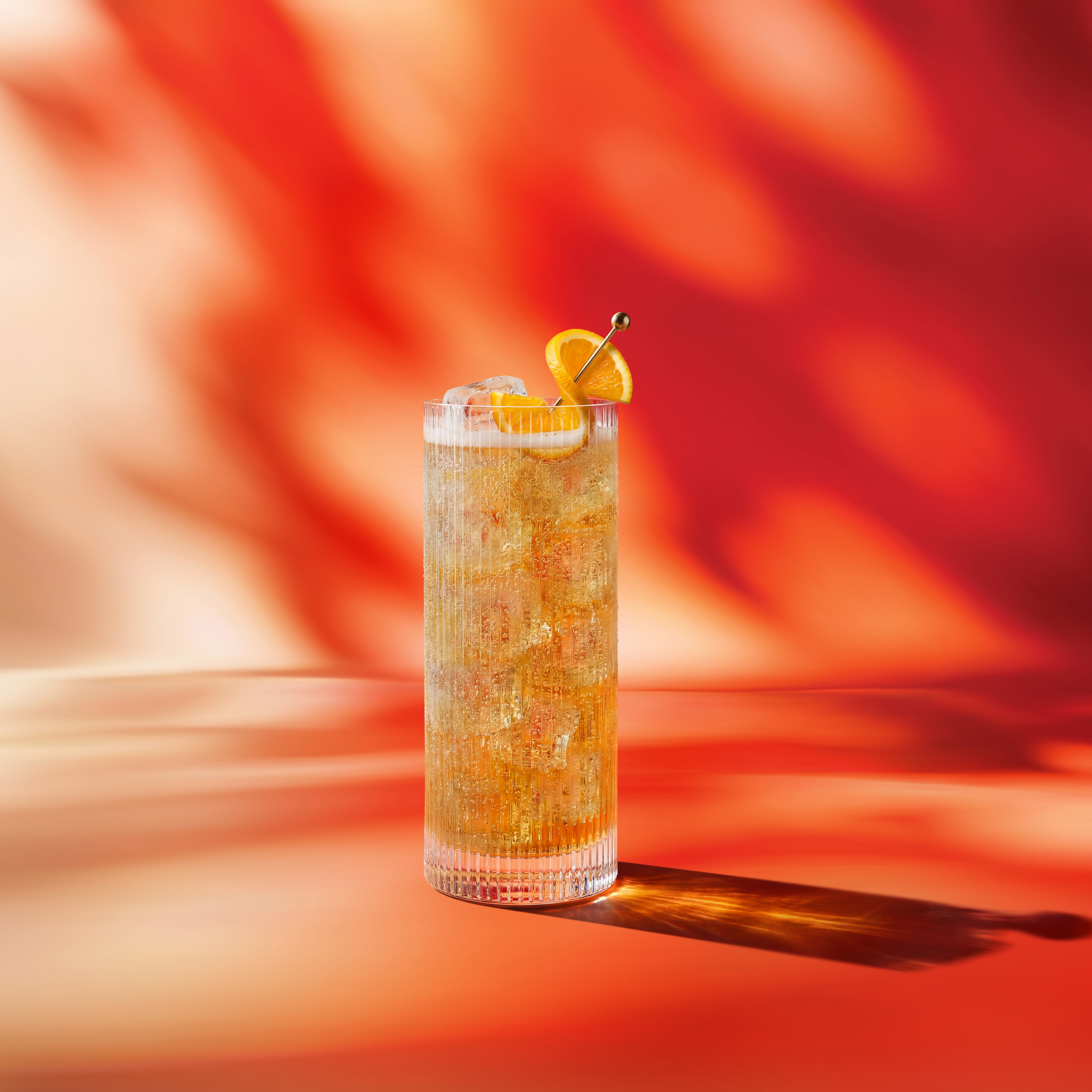 Un cocktail „highball” decorat cu o felie de portocală, preparat cu whisky Red Soul, pe un fundal cu pete roșii și albe.