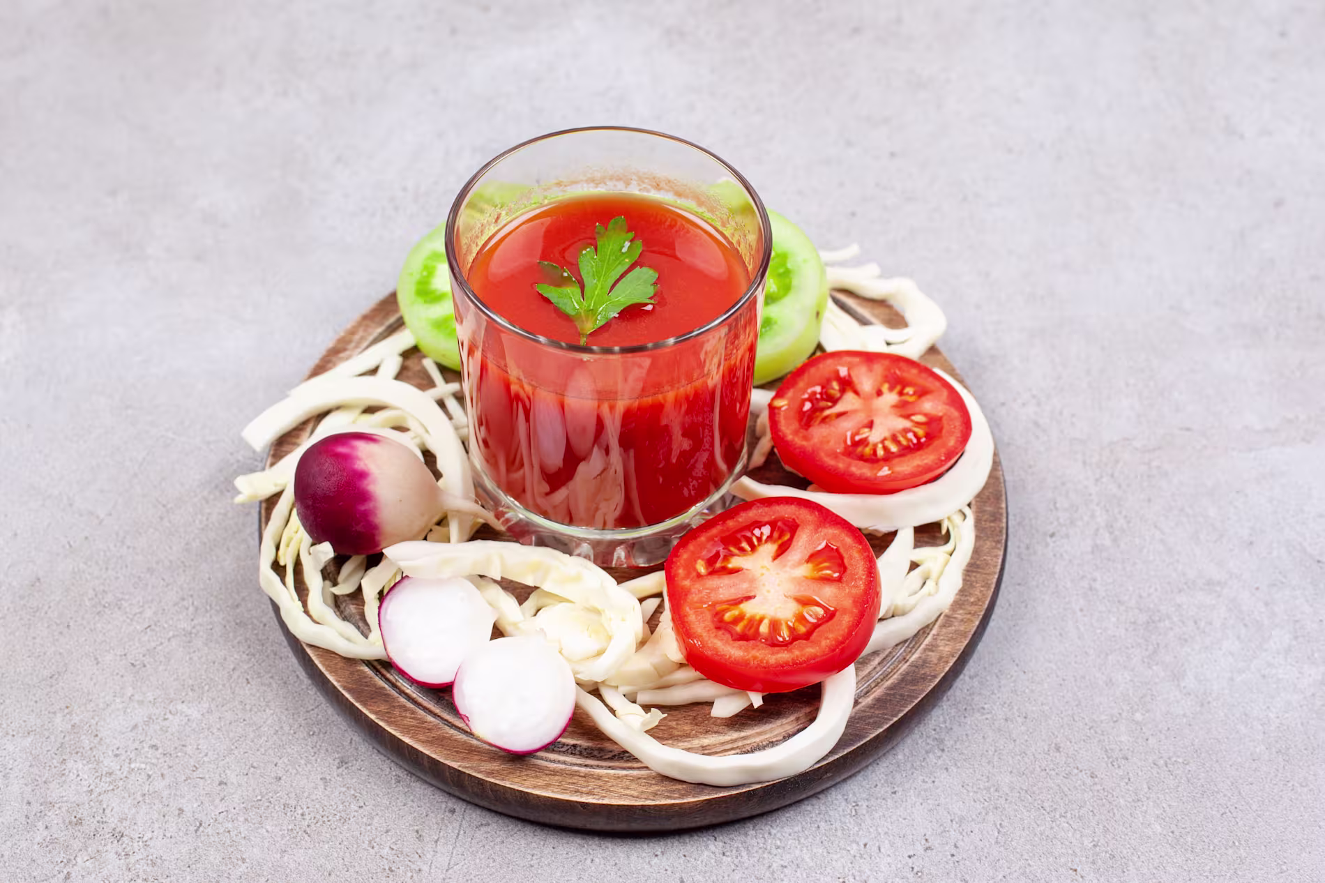 A glass of tomato juice garnished with parsley, surrounded by sliced tomatoes, radishes, cabbage, and a green tomato on a wooden plate.