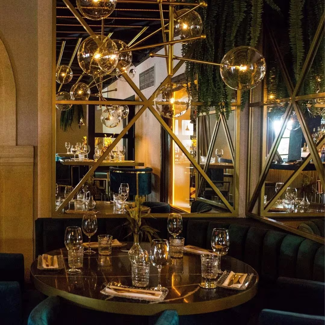 Espacio sofisticado con mesa decorada, rodeada de espejos y detalles en dorado. Las lámparas de vidrio aportan un toque moderno y elegante, ideal para una experiencia de alta gastronomía.