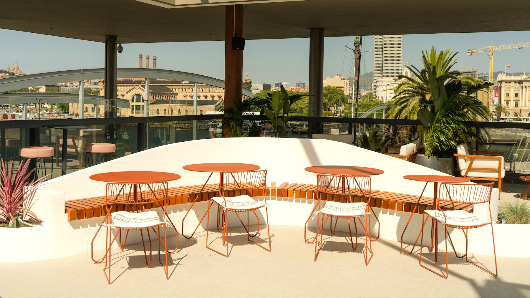 Terraza soleada con vistas al puerto de Barcelona, mesas naranjas, sillas de diseño y palmeras que completan un rincón moderno y relajado.
