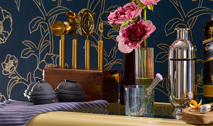 A bar setup with cocktail tools, a shaker, bottles, a glass with swizzle sticks, a vase with pink flowers, and two black tea infusers, set against a dark floral wallpaper.