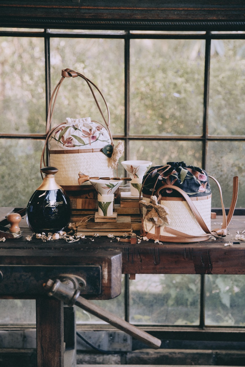 Two woven baskets, cups with botanical prints, a dark glass bottle, and small wooden items are arranged on a wooden table in front of a large window.