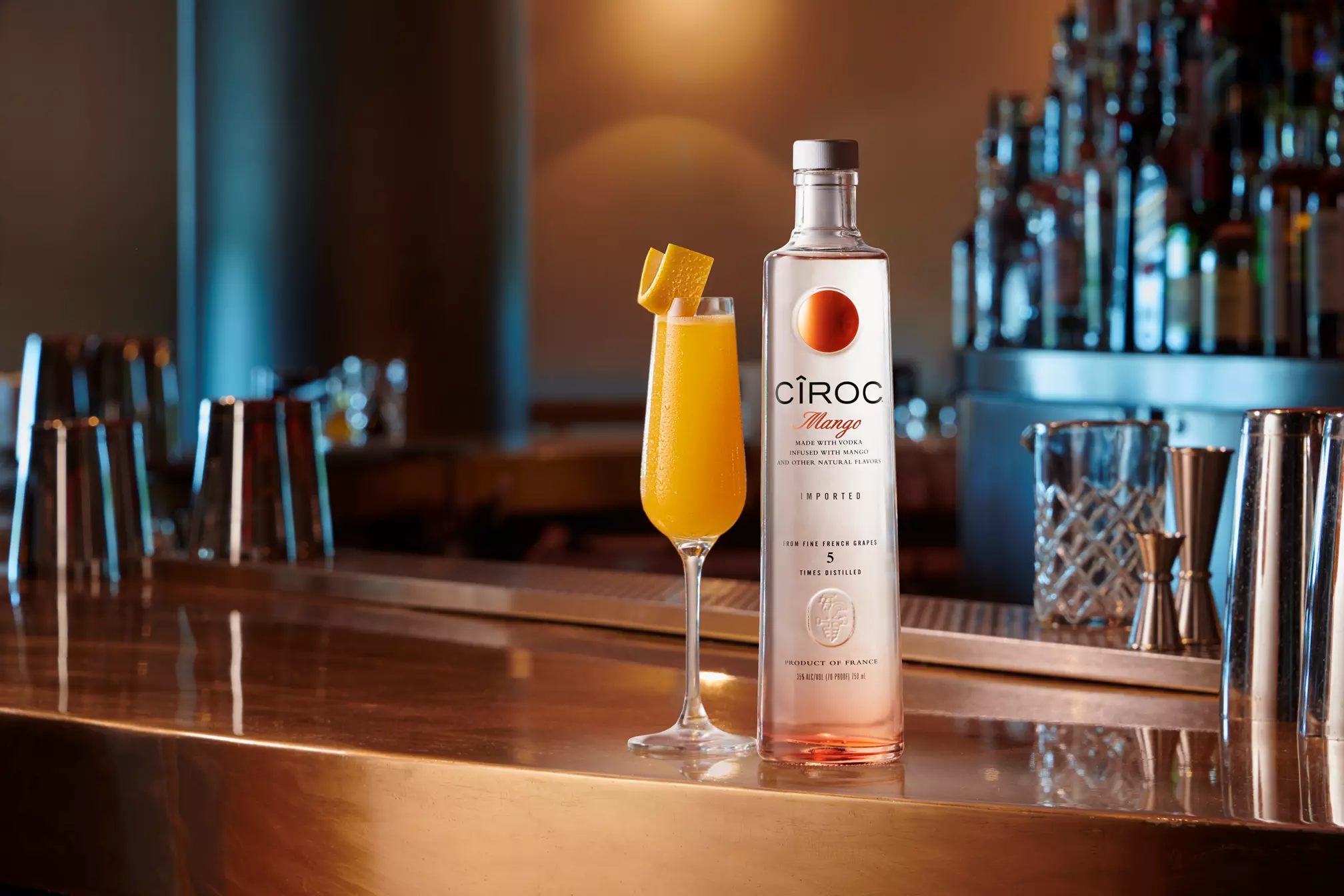 A bottle of Ciroc Mango vodka stands next to a tall glass of orange cocktail garnished with a citrus peel on a polished bar counter.