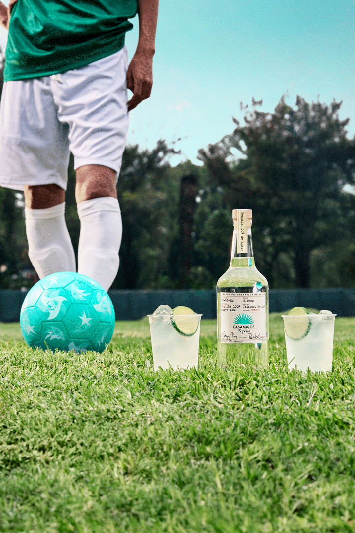 A soccer player stands on grass next to a blue soccer ball, with a bottle of tequila and two cocktails garnished with lime in the foreground.