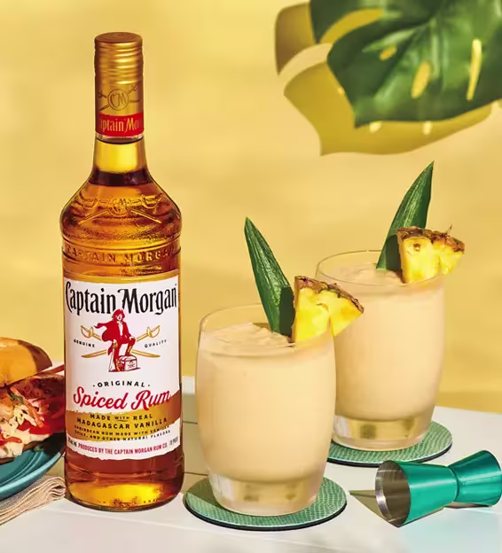 A bottle of Captain Morgan Original Spiced Rum next to two tropical cocktails garnished with pineapple and leaves on a table.