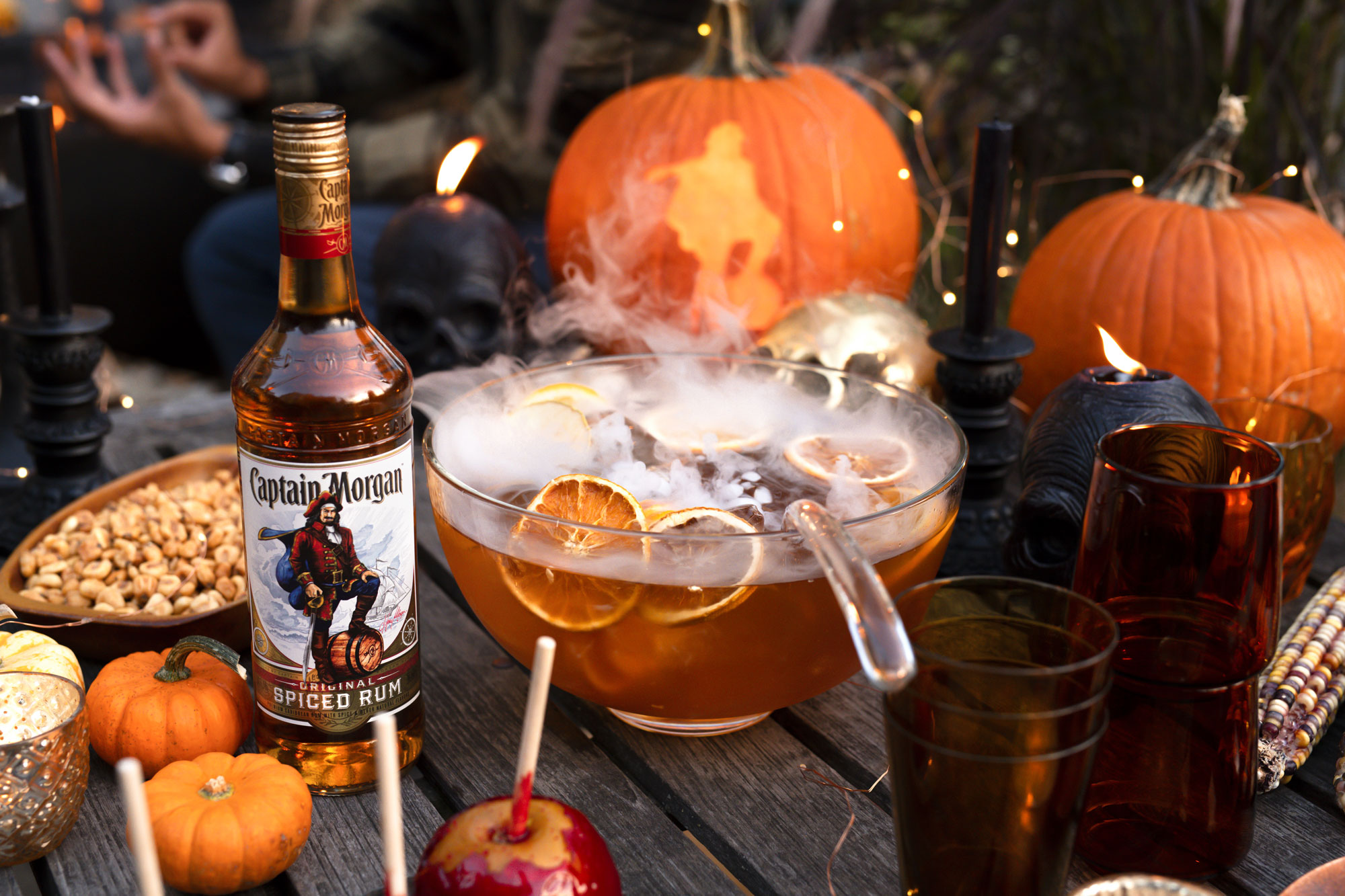 A bottle of Captain Morgan Spiced Rum next to a smoking punch bowl, glasses, pumpkins, skull decorations, and snacks on a Halloween-themed table.