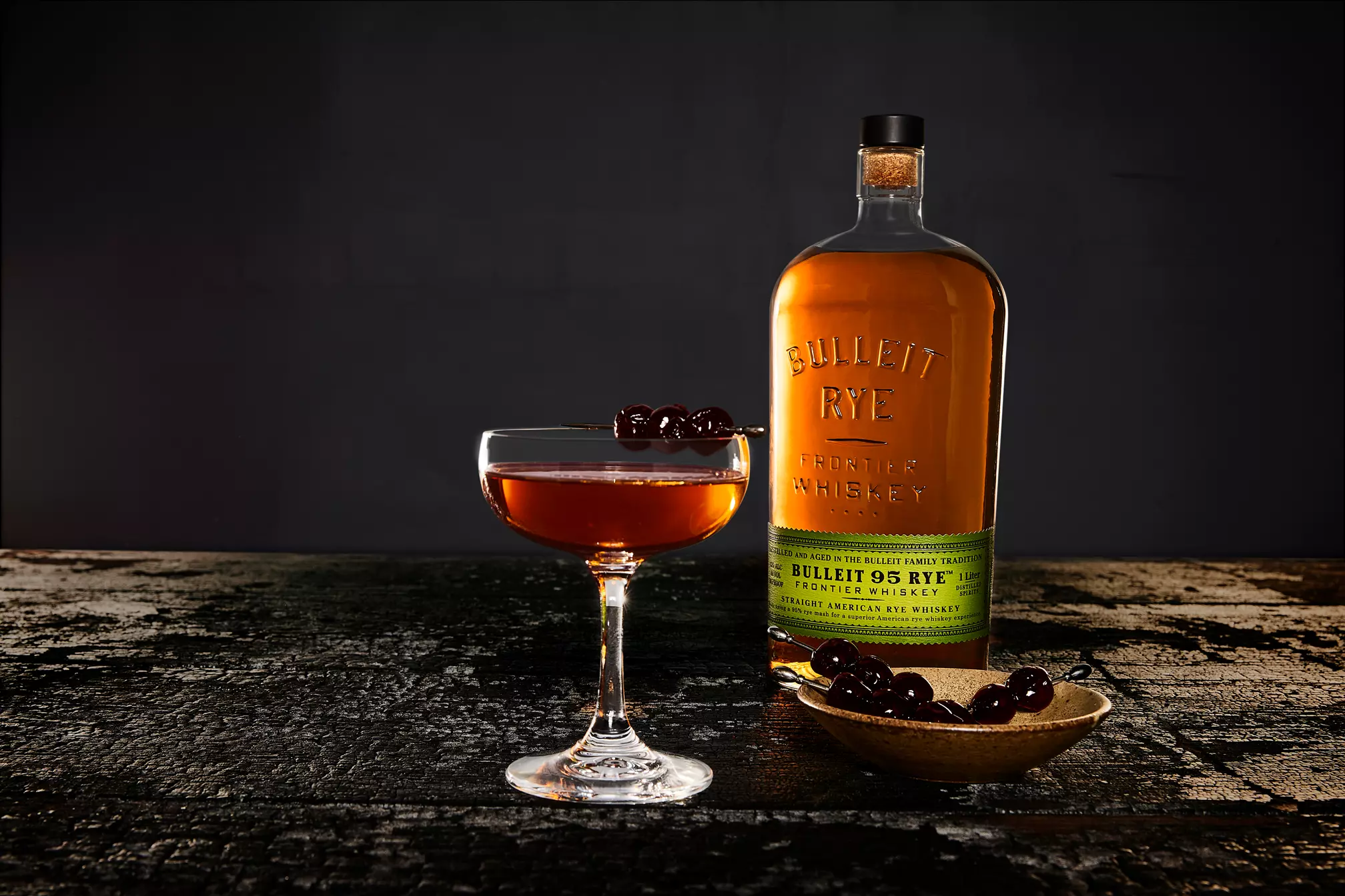 A bottle of Bulleit Rye whiskey stands next to a cocktail glass filled with a dark drink and garnished with cherries; extra cherries sit in a small bowl nearby.